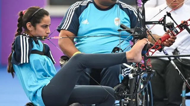 A foto mostra uma mulher usando uma camiseta azul,quantos saques posso fazer na greenbetspele clara e cabelos cumpridos presosquantos saques posso fazer na greenbetsuma trança. Ela está sentada, operando o arco com a perna direita. Ela não tem os braços.