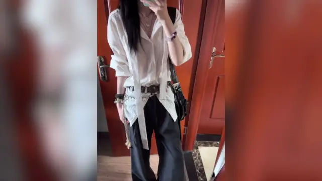 Kırmızı tuğla arka planlı bir aynanın önünde selfie çeken, siyah pantolon ve uzun püsküllü beyaz gömlek giymiş kadın.