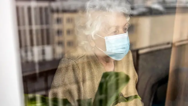 Mulher idosa de máscara olhando pelo vidro de um janela
