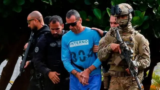 Detento algemado e com uniforme azul é escoltado por agentes de segurança fortemente armados