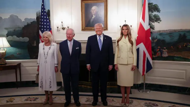 Kraliçe Camilla, Kral Charles, Donald Trump ve Melania Trump iki ülke bayrağı önünde ayakta poz veriyor. Erkekler koyu takım elbise, kadınlar açık renk giyinmiş, ve duvarda george washington tablosu var