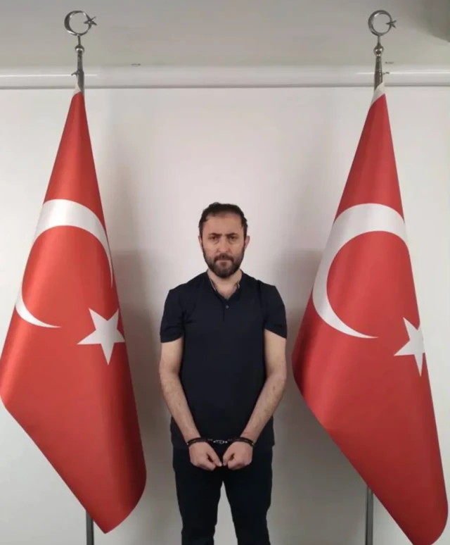Elleri önden kelepçeli siyah giysili bir adam. İki ynaında türk bayrakları var.