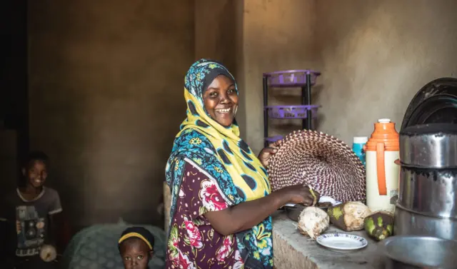 Une femme dans ce qui ressemble àune cuisine, sourit à la caméra avec deux enfants derrière elle.