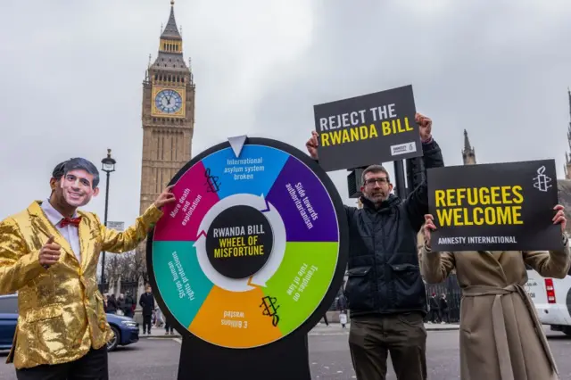 Un acteur portant un masque de Rishi Sunak et déguisé en animateur de jeux vidéo fait tourner une "roue de l'infortune" sur Parliament Square lors d'une manifestation organisée par Amnesty International UK pour souligner les graves conséquences négatives du projet de loi du gouvernement sur la sécurité du Rwanda, le 11 mars 2024 à Londres, au Royaume-Uni. 