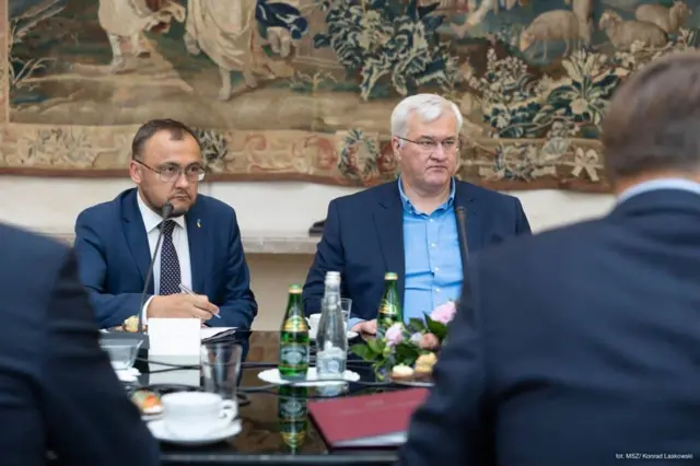 Офіційна зустріч: за столом сидять Боднар і Сибіга, перед ними розміщені мікрофони, документи, пляшки з водою та чашки. На столі також видно невелику квіткову композицію. На задньому плані висить великий декоративний гобелен із рослинними та фігурними мотивами.