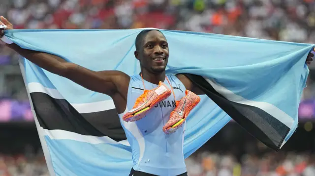 Letsile Tebogo, vêtu d'une veste de course bleue du Botswana, tient un drapeau du Botswana, les bras tendus et ses chaussures de course orange autour du cou.