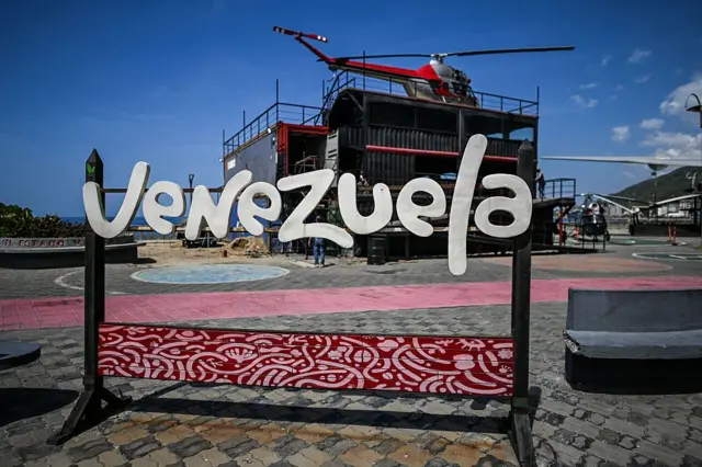 লোহার একটি স্ট্যান্ডে ইংরেজিতে ভেনেজুয়েলা লেখা, পেছনে একটি মাচার মতো কাঠামোর ওপর ও পাশে কয়েকটি হেলিকপ্টার রাখা 