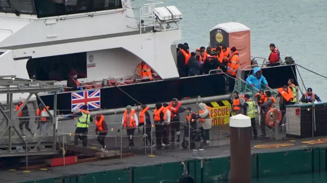 オレンジ色の救命胴衣を着た20人以上の人たちが、イギリス国旗がついた船から降ろされ、埠頭(ふとう)に立っている