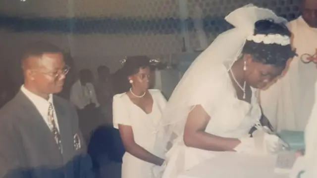 Mwanamume aliyevalia suti ya kijivu anamtizama bibi harusi aliyevalia nguo nyeupe akitia sahihi kwenye cheti. Mwanamke mwingine aliyevalia nguo nyeupe pia amesimama akimwangalia bibi harusi. 