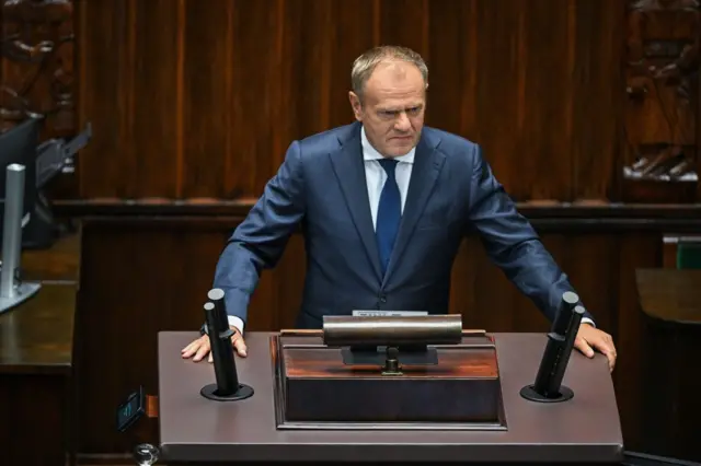 El primer ministro polaco, Donald Tusk, vestido con traje de chaqueta y gesto serio, hablando en el estrado del parlamento con sus manos extendidas sobre él. Todo el fondo y el estrado son de madera oscura.