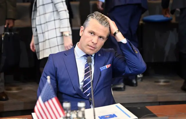 Pete Hegseth saçını düzeltiyor. Takım elbise giymiş. Önünde Amerikan bayrağı var.