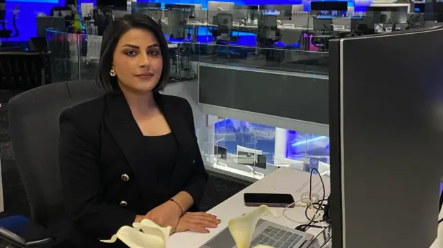 A woman with dark hair sitting at a desk in a TV newsroom looks at the camera