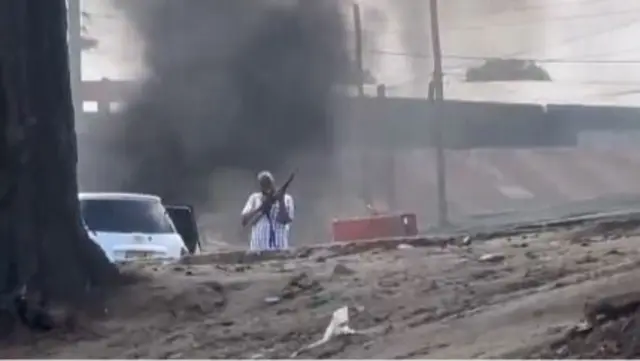 Mwanamume aliyevaa mavazi ya kiraia ameshikilia bunduki, amesimama kando ya gari huku moshi mweusi ukifuka nyuma yake