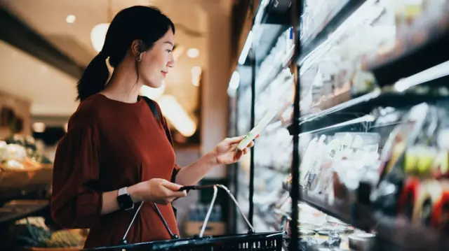  یوې ښځې په سوپرمارکېټ کې هغه محصول ګوري چې نیولی یې دی، هغه د یخچال څانګه کې ولاړه ده. هغې نسواري جامه اغوستې او په یوه لاس کې یې ټوکرۍ نیولې ده.