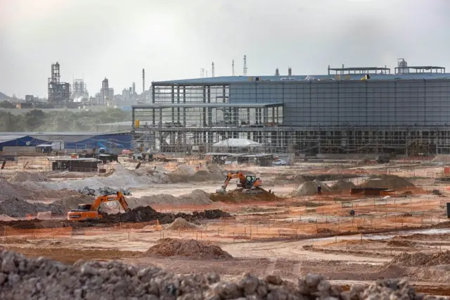 Obra da construção da fábrica da BYD em Camaçari com escavadeiras, pilhas de materiais e estruturas metálicas ao fundo