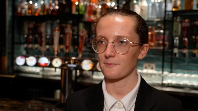 Chloe aparece en un bar. Se ven los surtidores de cerveza detrás de ella. Lleva una chaqueta negra y gafas redondas. Lleva el pelo recogido.