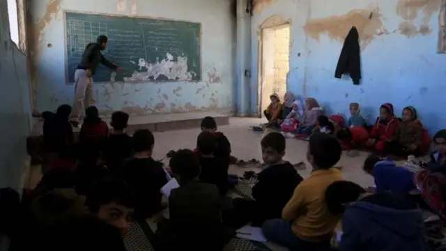 أطفال من البنين والبنات وأماهم معلم يشرح على سبورة في مدرسة في سوريا