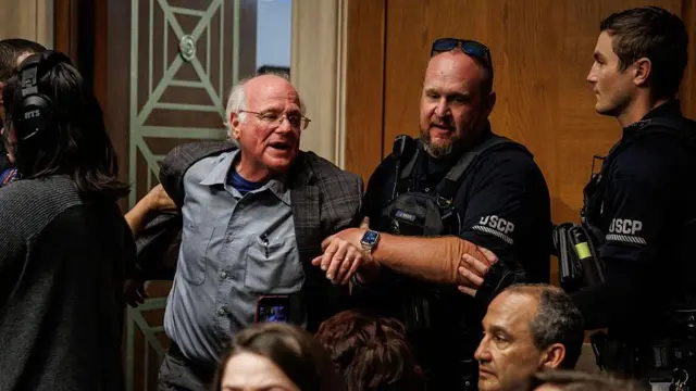 Двое мужчин в форме выводят под руки пожилого человека в серой рубашке и пиджаке и в очках