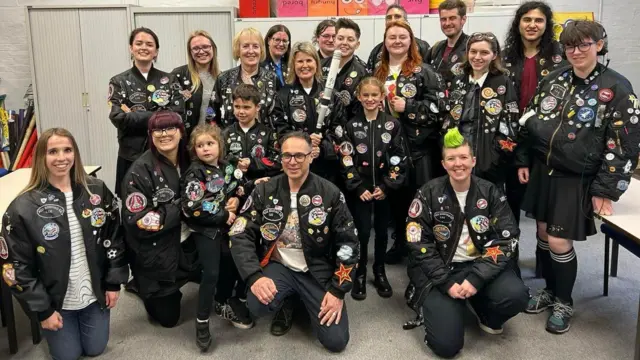 A group of fans of different genders and ages all wearing Ace's iconic jacket with Sophie stood in the middle holding a baseball bat.