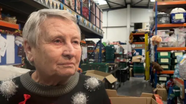 Mary lleva un jersey navideño y está de pie en un almacén. Detrás de ella se ven muchas cajas llenas de donaciones apiladas en los estantes, así como cajas de cartón vacías esperando a ser llenadas.