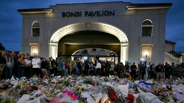 На заднем плане видно большое арочное здание с надписью Bondi Pavilion. На переднем плане — большая композиция из цветов, израильских флагов и других предметов, уложенная на полу перед собравшейся толпой людей.