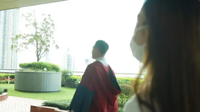一名身穿學位袍的男子站在室外草坪邊，望向遠方的高樓景色，前景中有另一名戴口罩的女子部分入鏡。
