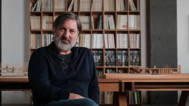 Hombre sentado en un estudio con estantes llenos de libros y maquetas arquitectónicas de fondo.