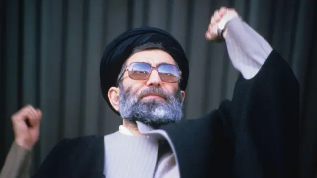 Ayatollah Khamenei making a speech in 1986. He is wearing a black robe with grey shirt and black head covering. His fists are in the air.