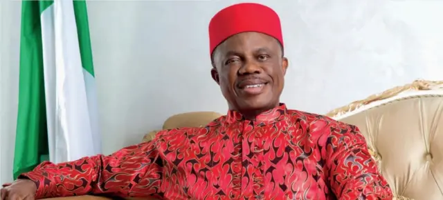 Obiano yi uwe na-acha uhie uhie nwere ntụpọ ojii, kpuru okpu uhie, nọrọ n'oche ebe ọkọlọtọ Naịjirịa dị ya n'azụ.