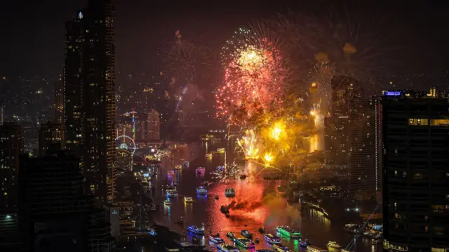 暗いビル群の間を流れる川の上で、オレンジと黄色の花火が上がっている。川の上に並ぶ船は緑や紫、青などにライトアップされている