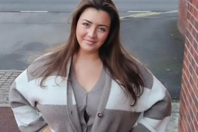 Melissa Ashcroft wearing a grey and white striped cardigan and blue jeans. She has straight brown hair and is standing with her hands on her hips. 