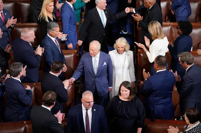 У великій залі з парламентськими лавами людина в темному формальному костюмі йде проходом між рядами сидінь. Поруч — ще одна людина в світлому формальному вбранні. Довкола стоять і сидять присутні в діловому одязі, багато з них аплодують. Атмосфера урочиста, офіційна, зосереджена на центральних фігурах у проході.