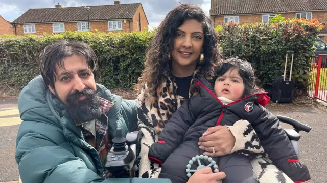Un hombre y una mujer sostienen a un niño pequeño. El hombre tiene el pelo corto y oscuro, barba y lleva una chaqueta acolchada de color verde grisáceo. La mujer tiene el pelo largo, oscuro y ondulado, y lleva una bufanda con estampado de leopardo. El niño pequeño tiene el pelo oscuro y lleva un mono acolchado gris oscuro con cremallera.