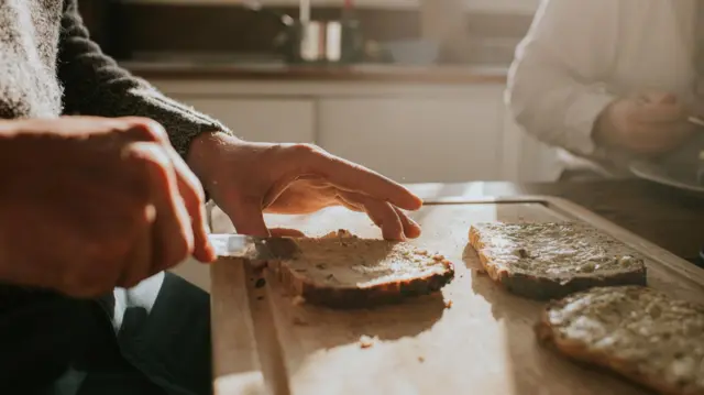 لقطة مقرّبة لرجل يدهن الزبدة على خبز أسمر محمص قليلاً في مشهد إفطار صباحي. مشهد بسيط لتحضير وجبة في صباح مشمس.