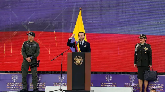 De la Espriella faz seu discurso de posse em um palco em frente a uma tela exibindo a bandeira colombiana e acompanhado por dois representantes da força pública, um homem e uma mulher.