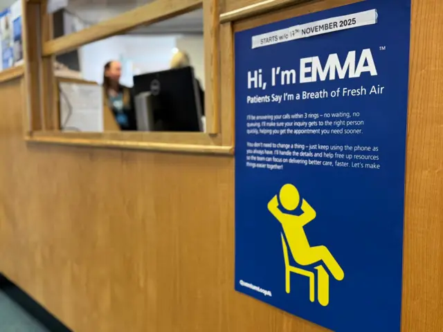 Un póster azul con una figura amarilla sentada en una silla. El póster tiene un texto blanco grande que dice "Hola, soy Emma". Está colocado en una pared de madera. Al fondo se ve un mostrador de recepción y dos personas.