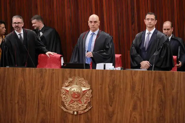 Alexandre de Moraes em pé em frente a mesa do TSE, rodeado por ministros e assessores; ele tem olhar atento e sério 