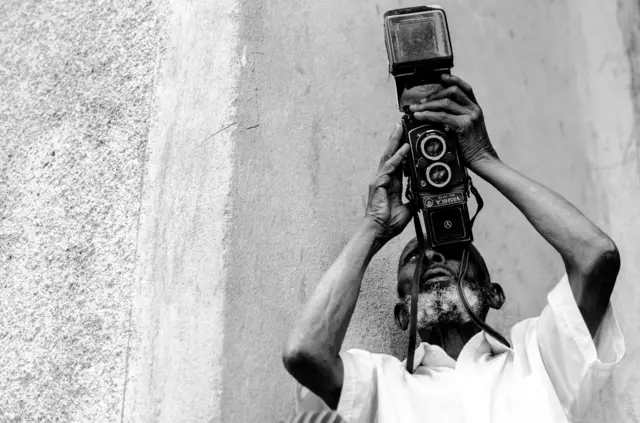 የዐዲስ አያት፣ አቶ ሰፊው ካሜራውን ወደ ላይ አድርገው ሰዎችን ፎቶ ሲያነሱ 