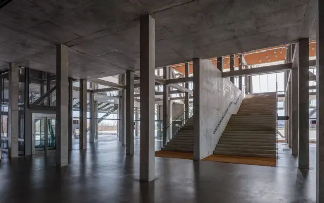Interior amplio con estructura de hormigón y metal, columnas expuestas y una escalera central iluminada por luz natural.