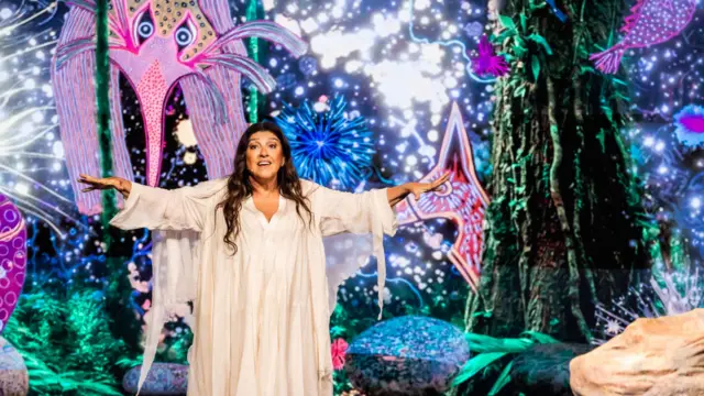 Regina Casé veste uma longa túnica branca e aparece no centro do palco com os braços abertos. Ao fundo, projeções coloridas de criaturas marinhas e formas orgânicas criam um cenário que remete aos primórdios da vida na Terra, complementado por elementos cenográficos que simulam rochas e vegetação.