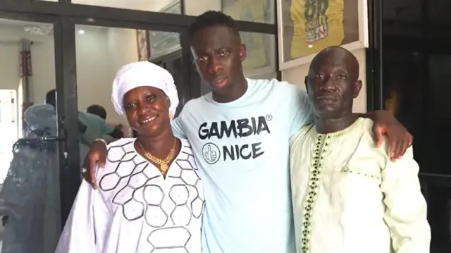 Yankuba Minteh, vêtu d'un t-shirt portant le message « Gambia Nice » et une image caricaturale représentant un pouce levé, enlace sa mère Jassey et son père Buhari, qui se tiennent respectivement à gauche et à droite de leur fils, face à l'appareil photo. Jassey porte un voile blanc, un collier en or et une robe blanche traditionnelle, tandis que Buhari est vêtu d'une robe vert clair.
