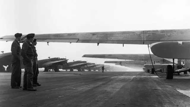An avenue of Hamilcar gliders (right) and RAF Halifax towing aircraft (left) on 24 March 1945. The black and white image shows three aircrew with their hands in their pockets looking towards the gliders.