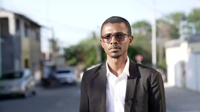 Homem negro com óculos escuros, terno preto e quipá na cabeça em bairro periférico