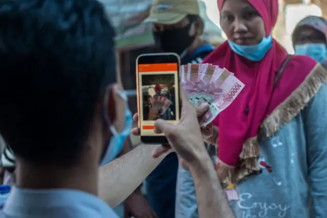 Seorang petugas memotret uang kertas rupiah Indonesia bersama penerimanya saat warga, yang mata pencahariannya terdampak pandemi Covid-19, menerima bantuan tunai dari pemerintah di Surabaya pada 19 Juli 2021.