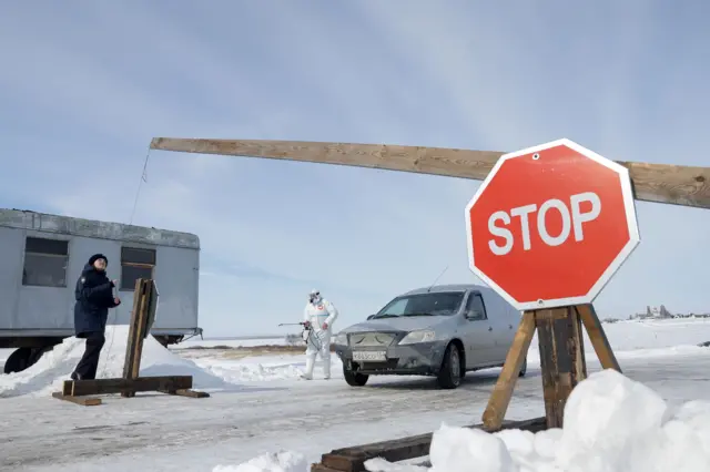 Контроль на въезде в село Козиха в Новосибирской области, где введен карантин для местного скота. На фото полицейская поднимает шлагбаум для машины, а рядом стоит человек в защитном костюме, который обрабатывает машины.
