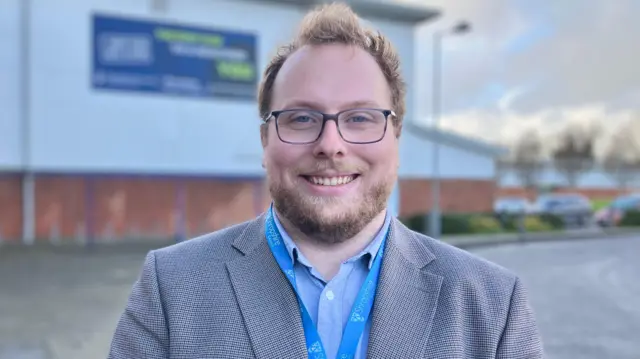 Un hombre de pelo rubio y barba, vestido con una chaqueta gris y un cordón azul, de pie frente a un edificio revestido de blanco con una base de ladrillo.