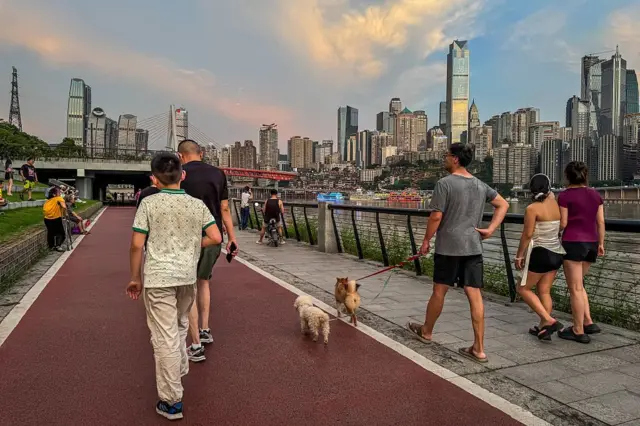 Ludzie wyprowadzają psy, ćwiczą i spacerują wzdłuż nadrzecznej ścieżki dla pieszych, w tle widać panoramę miasta i most wantowy o zachodzie słońca