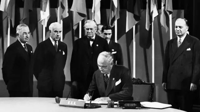 Чоловік підписує документ за столом. Позаду стоять четверо інших чоловіків