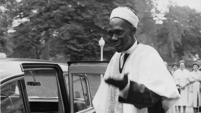 Tafawa Balewa, Olootu ijọba Naijiria tẹlẹ. O we lawani le ori agbada funfun to wọ, o n rẹrin-in bo ṣe duro lẹgbẹẹ ọkọ rẹ, o si nawọ siwaju. Awọn obinrin oyinbo kan han lẹyin rẹ ninu fọto yii.