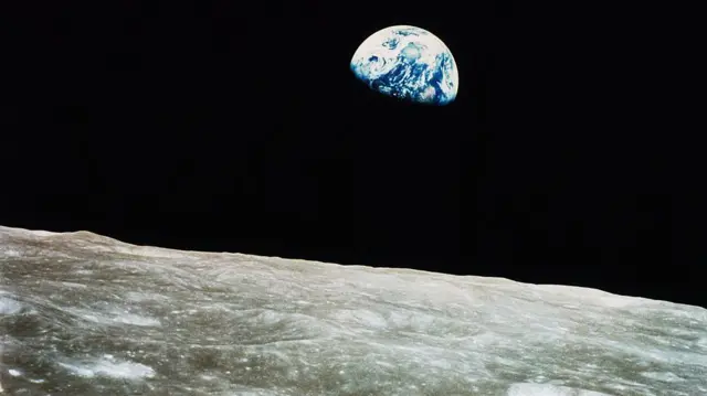 Em primeiro plano, vê-se uma pequena porção da superfície lunar prateada, com uma visão parcial da Terra como uma bola azul com espirais brancas "ascendendo" em um "céu" completamente preto.